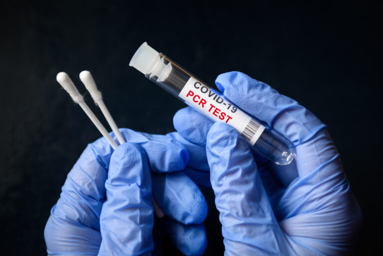 Gloved hands holding a tube labeled COVID-19 PCR test and swabs.